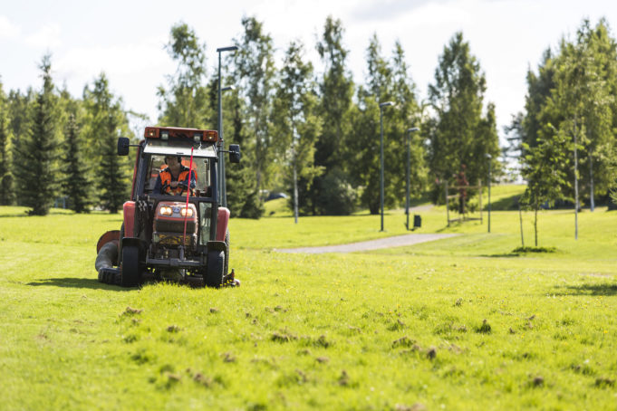 Pieni punainen traktori leikkaa nurmikkoa kesällä Penttilänpuistossa.