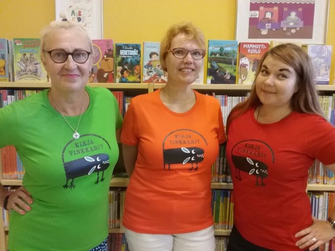 Kirjaston kirjavinkkarit poseeraavat kirjahyllyn edessä pukeutuneina Kirjavinkkari-paitoihin.