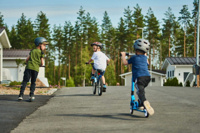 Kolme leikki-ikäistä poikaa ajavat polkupyörillä ja potkulaudalla Harjuniityn asuinalueen pihakadulla kesäpäivänä.