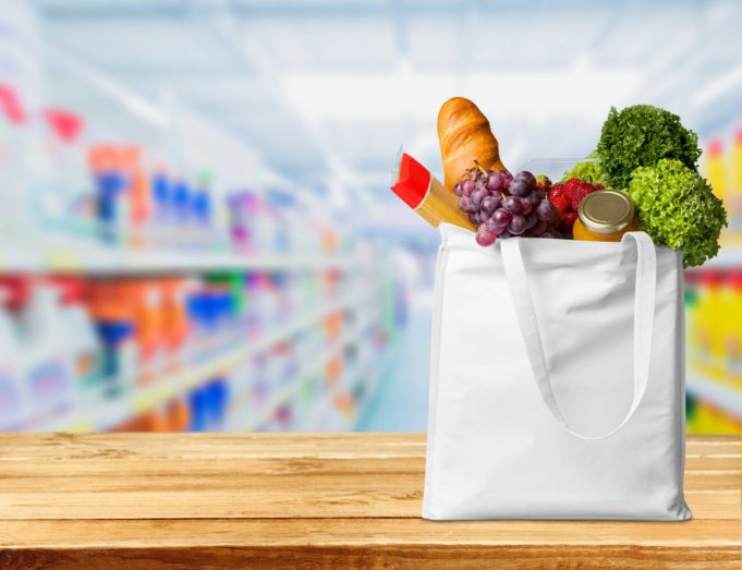 Valkoinen ostoskassi pöydällä täynnä elintarvikkeita: vihanneksia, leipää, spagettia ja säilykkeitä.