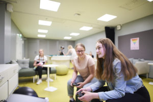 Nuorisotila Nuksussa kaksi nuorta pelaa etualalla konsolipeliä. Taustalla nuoria pelaa biljardia ja istuu sohvalla.