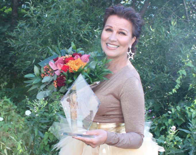 Arja Koriseva hymyilee kädessään lasinen Iskelmä Finlandia-palkinto ja kukkakimppu, jossa on punaisia kukkia.