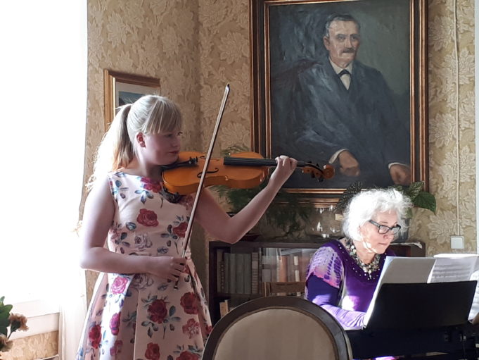 Nuori nainen soittaa viulua. Naisella on pitkät vaaleat poninhännälle sidotut hiukset ja kukallinen mekko. Hänen vieressään on pianoa soittava nainen. Pianistin takana seinällä on mieshenkilön muotokuva.