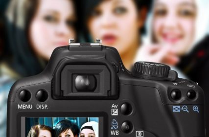 Kolme nuorta tummahiuksista naista katsoo kameraan, etualalla on digikamera, jonka ruudulla näkyy kolme kuvassa poseeraavat nuoret naiset.naista.