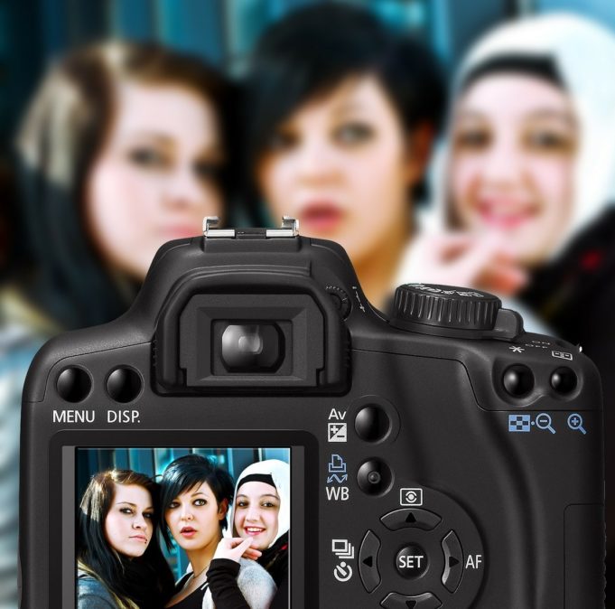 Kolme nuorta tummahiuksista naista katsoo kameraan, etualalla on digikamera, jonka ruudulla näkyy kolme kuvassa poseeraavat nuoret naiset.naista.