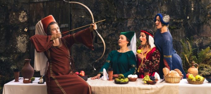 Neljä naishenkilöä ruokapöydän ympärillä. Yksi virittää jousta kohti taivasta, muut seuraavat hämmästynein katsein. Naiset ovat pukeutuneet keskiaikaisiin pukuihin.