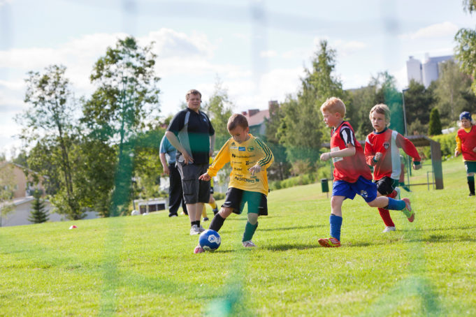 Kuvassa pelipaita-asuiset lapset pelaavat jalkapalloa nurmikentällä. Aikuinen katsoo taustalla peliä.