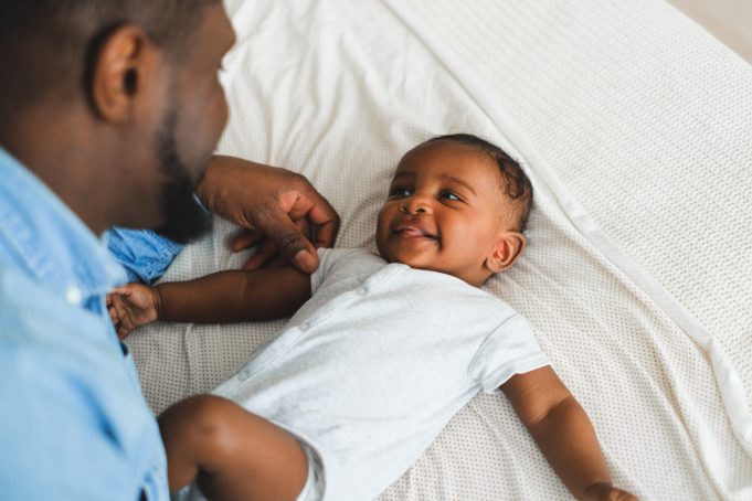 Kuvassa vauva makaa selällään ja mies on kumartuneena katomaan häntä. Vauva hymyilee miehelle. Vauvalla on päällään valkoinen, lyhythihainen body, miehellä sininen kauluspaita.