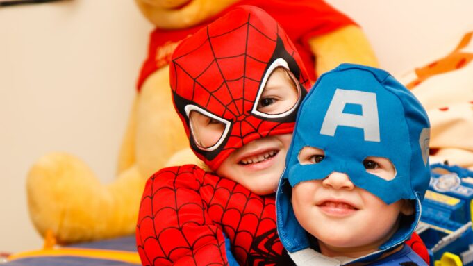 Kuvassa on kaksi lasta, joilla on supersankariasut päällään. Toisen lapsen päähineessä on iso A-kirjain. Lapset halaavat toisiaan. Taustalla näkyy iso Nalle Puh -pehmolelu.