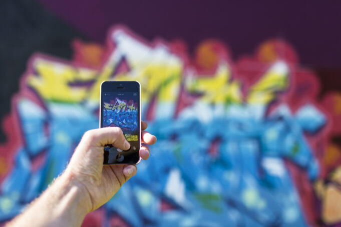 Kuvassa näkyy käsi, joka pitelee älypuhelinta ja on ottamassa valokuvaa takana näkyvästä värikkäästä graffitista.