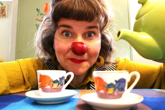 Punaiseen klovnin nenään sonnustautunut nainen pöydän ääressä, kuvan etualalla on kaksi värikästä kahvikuppia.