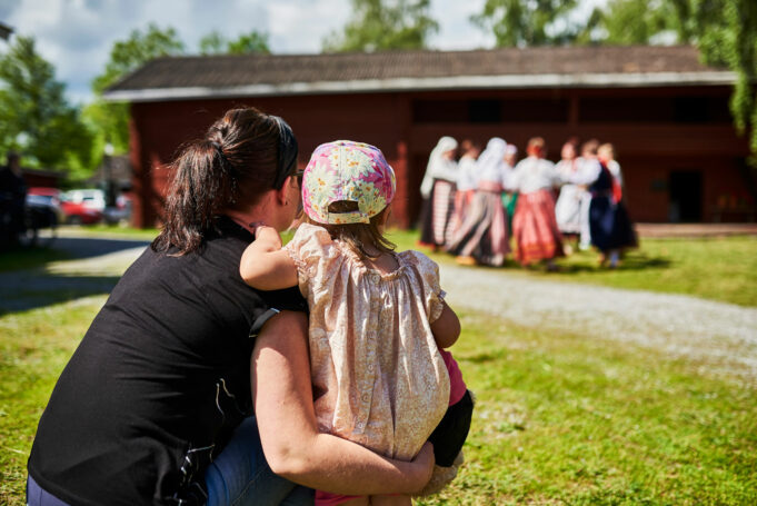 Nainen ja lapsi katsovat piirissä tanssivia kansallispukuisia esiintyjiä museon pihapiirissä.