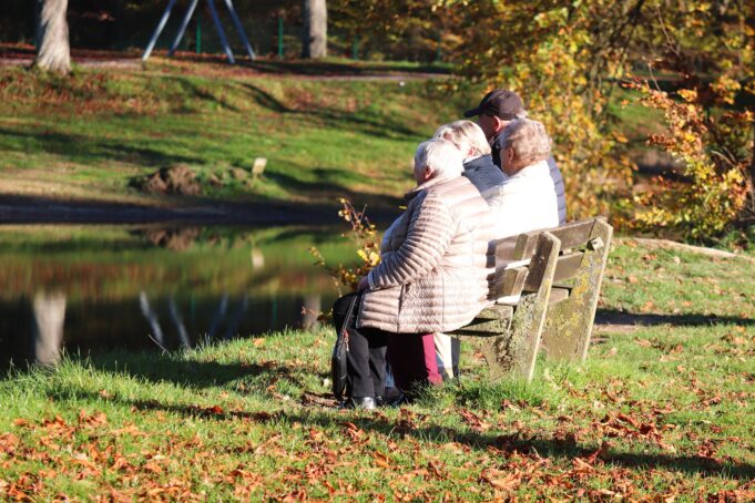 Neljä ikääntynyttä henkilöä istuu puiston penkillä katsellen lampea ja syksyistä maisemaa.